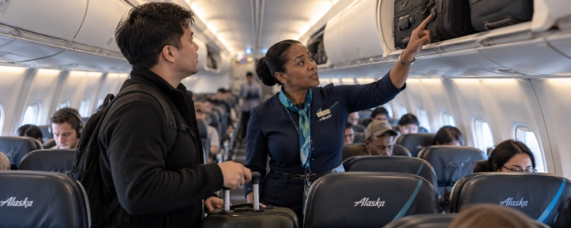 Flight attendant on an Alaska Airlines plane at Portland pointing to an overhead bin that is already full while a passenger holds a roller bag