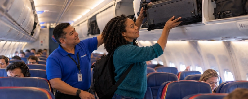 Woman on a Southwest plane at Dallas Love Field lifting a large gray roller bag into the overhead bin